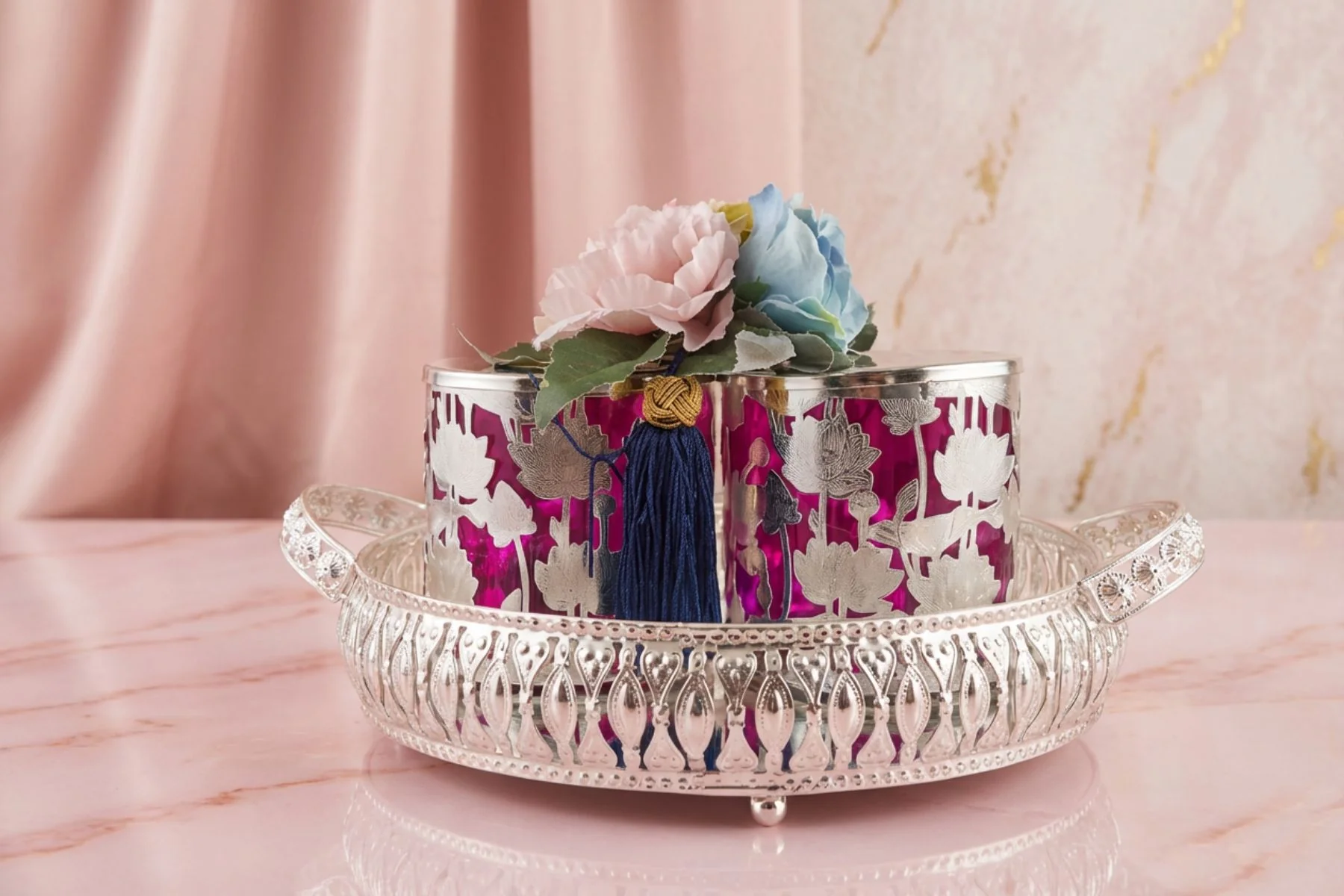 Ornate silver pedestal with magenta floral panels, pastel pink and blue flowers, and a blue tassel on a pink marble table surface.