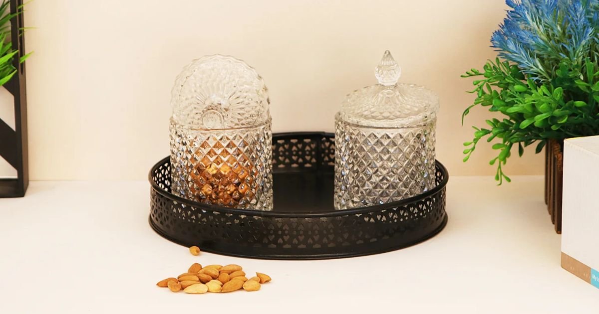 Two diamond-cut crystal jars with lids sit on a round black metal tray on a white surface, with almonds spilled nearby and green plants in the background.