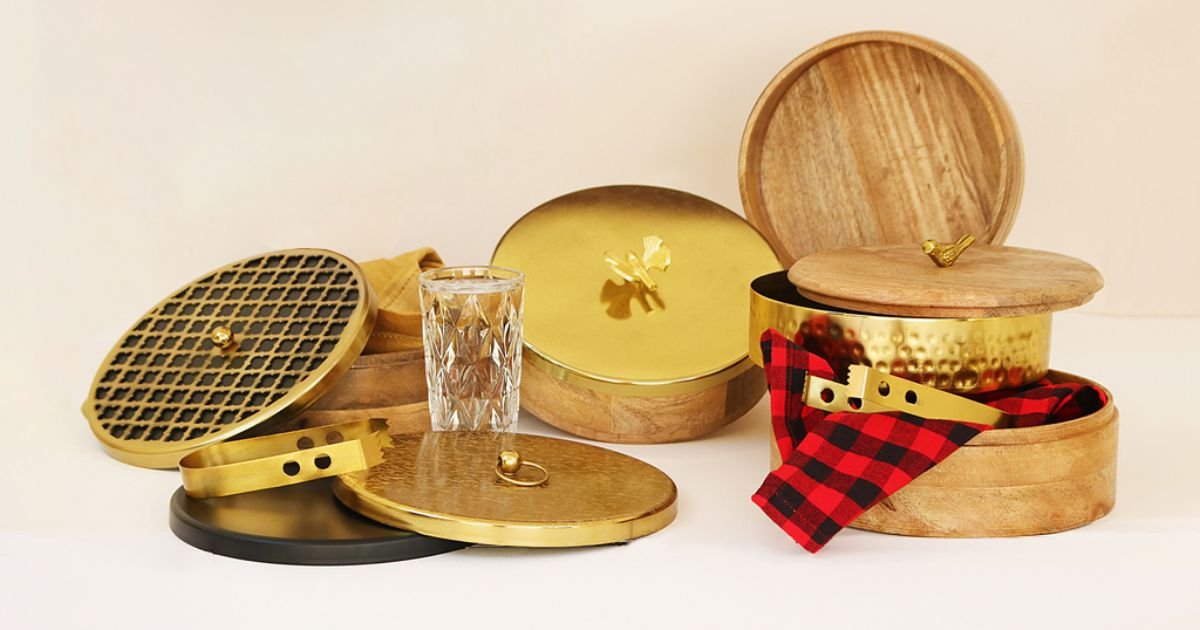 Assorted round brass and wooden containers with lids, arranged on a light surface, with a glass and a red plaid cloth nearby.