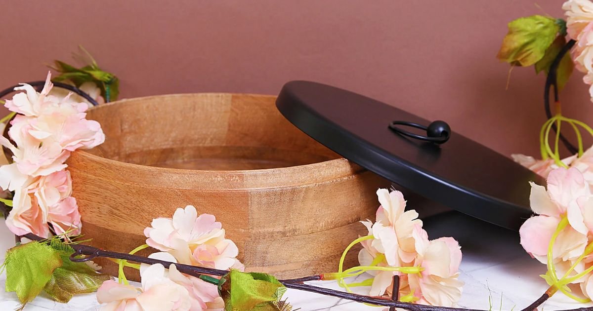 Open wooden tub with a black lid resting nearby, surrounded by pastel pink flowers and a floral garland, suggesting a spa scene.