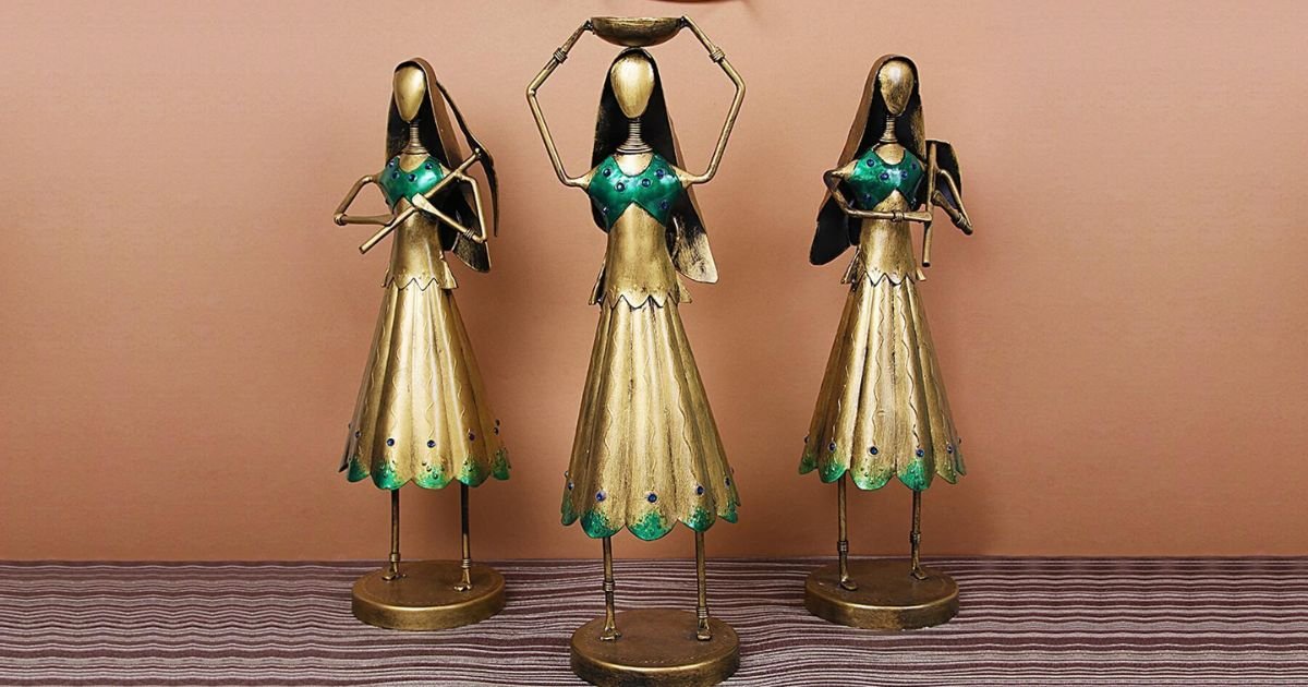 Three gold-toned metal figurines with green accents stand on a striped surface against a peach wall, posing as dancers with arms raised or crossed steadying their balance.