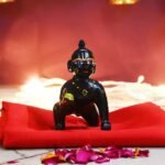 Small black Hanuman statue on a red cloth, part of a lit Hindu altar with golden vessels and candles nearby.