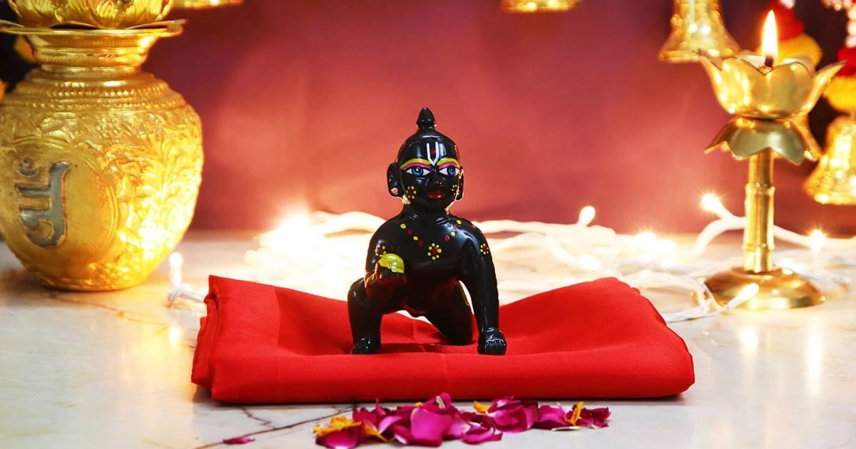 Small black Hanuman statue on a red cloth, part of a lit Hindu altar with golden vessels and candles nearby.