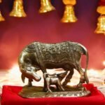 Bronze-toned statue of a cow and calf on a red cloth, displayed on a Hindu altar with lit lamps and gold ornaments in the background.
