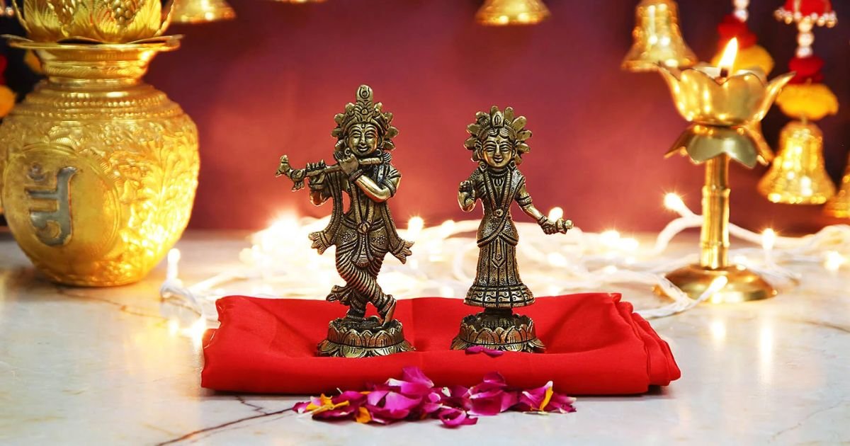 Two small brass figurines dancing on a red cloth, flanked by lit oil lamps and festive decor in a devotional altar setting.