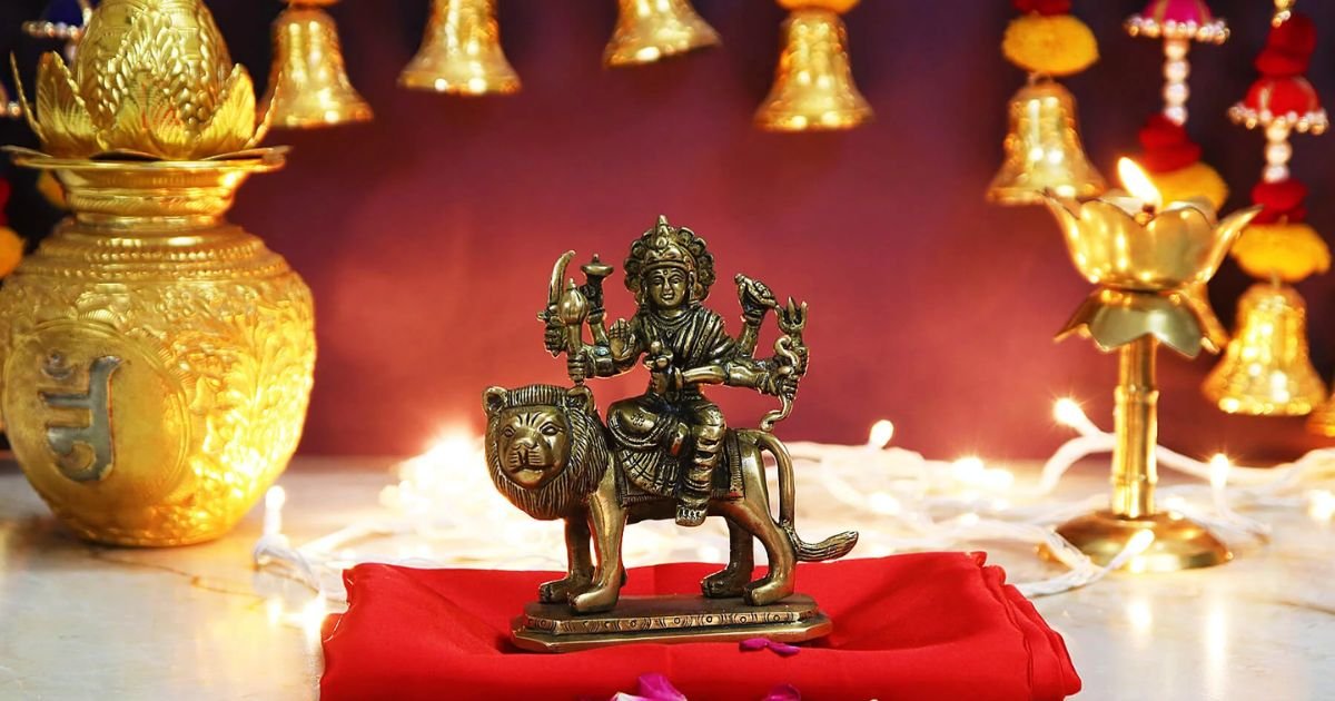Bronze statue of a Hindu goddess on a lion atop a red cloth, set in a shrine with glowing golden lamps.