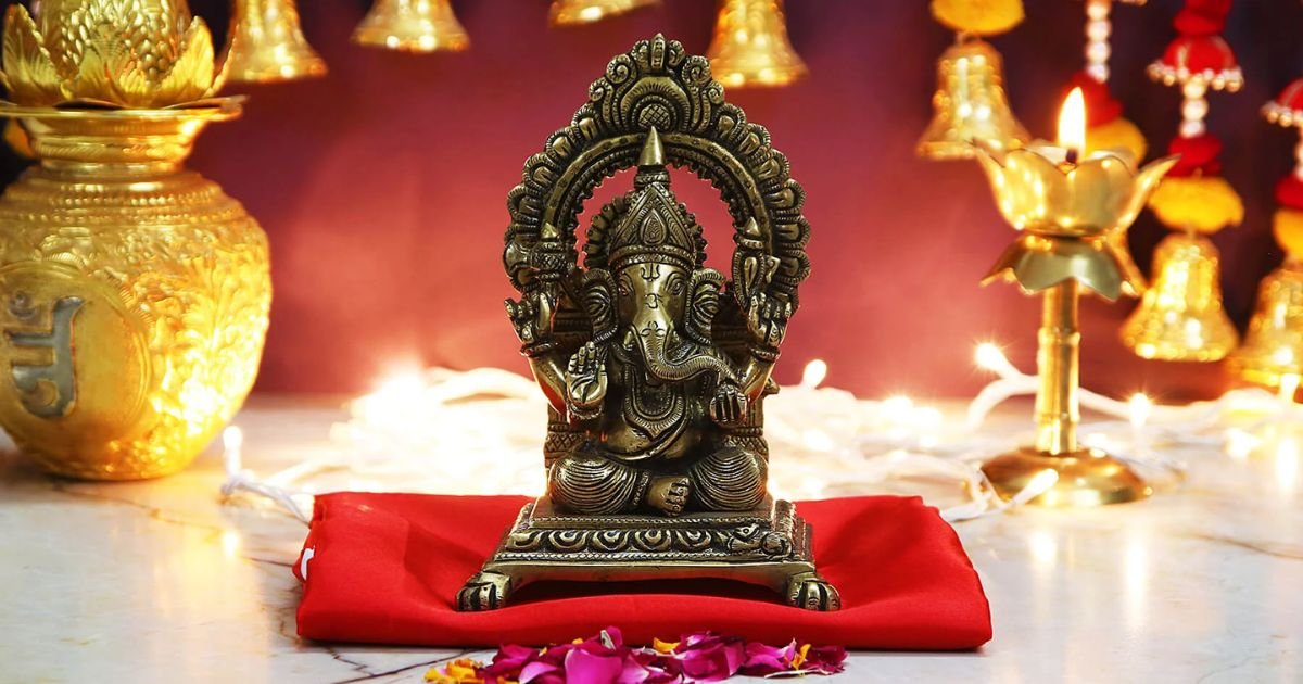 Bronze Ganesha statue seated on a red cushion in a lit shrine with gold decor and petals around it.