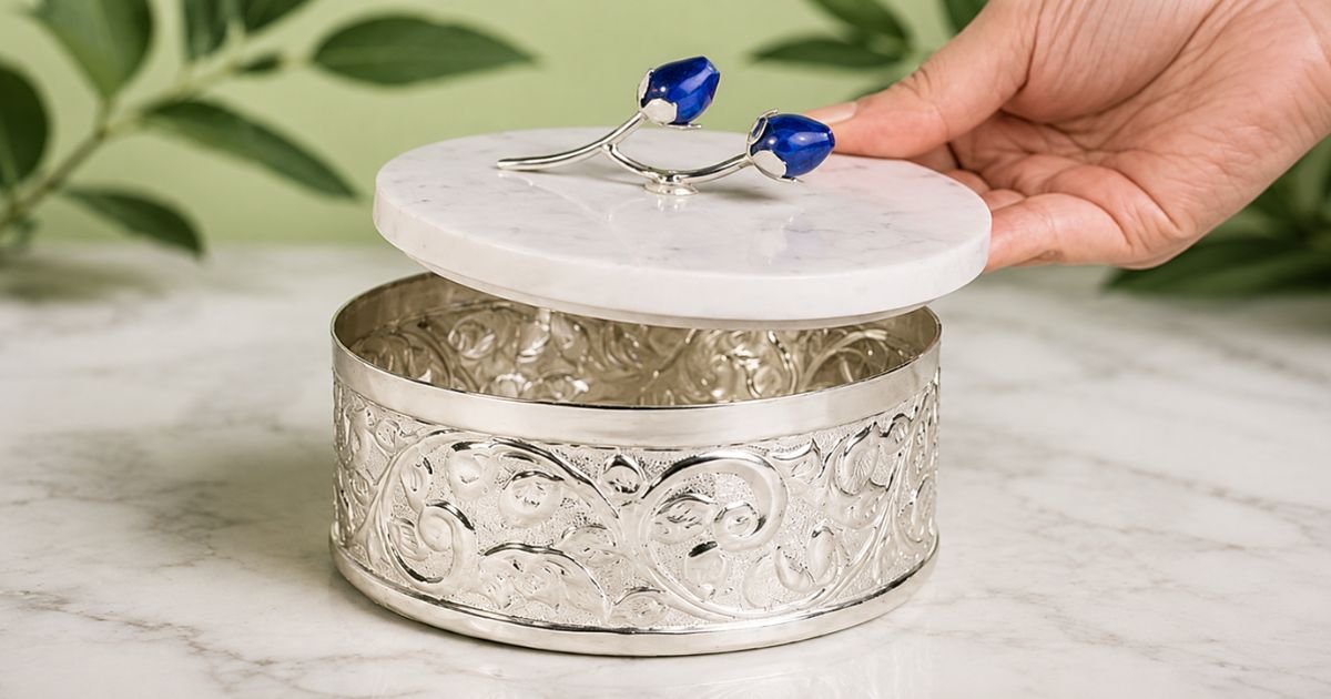 Ornate silver round jewelry box with a white marble lid, hand lifting the lid to reveal intricate scrollwork interior.