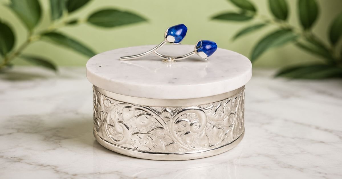 Round silver jewelry box with a white marble lid and intricate filigree base on a marble surface.
