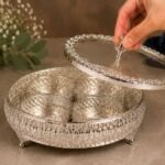 Hand lifting the lid of an ornate silver serving stand that holds four small cups inside.