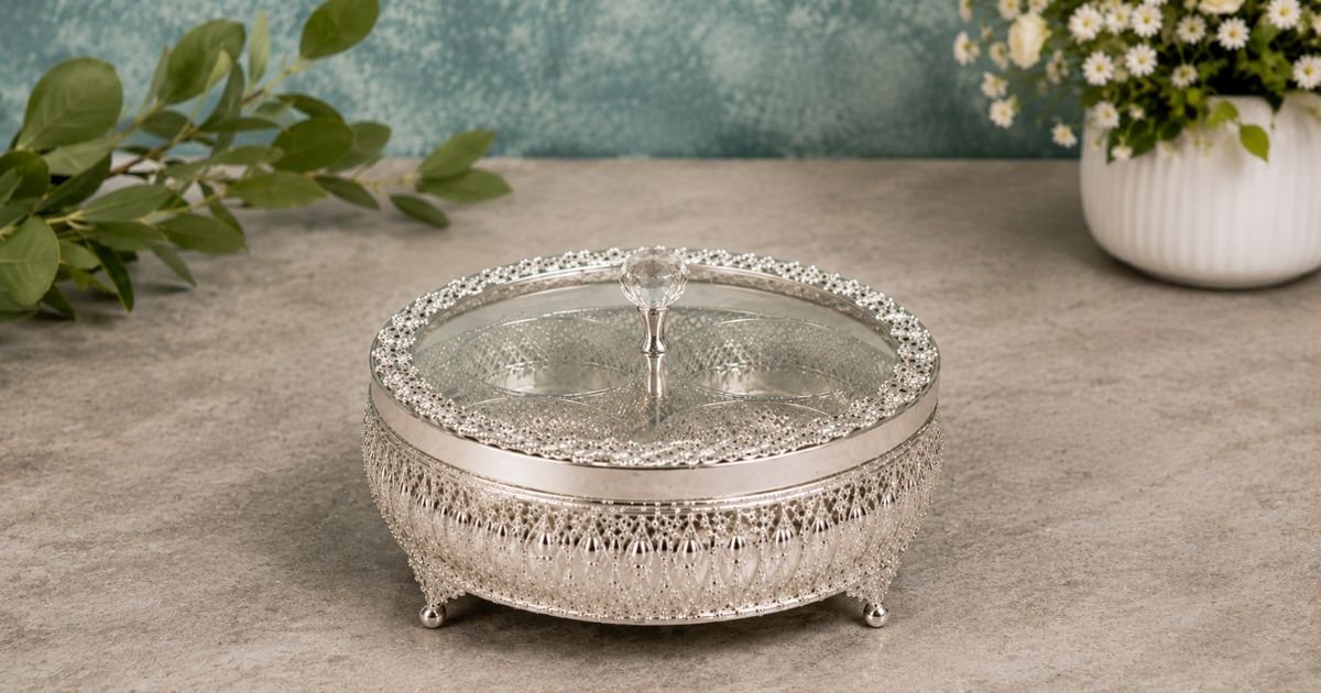 Ornate silver round trinket box with a glass lid and beaded filigree trim on short feet at a stone surface edge.