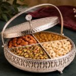 Ornate silver serving box with four compartments filled with almonds, pistachios, cashews, and raisins on a dark table surface.