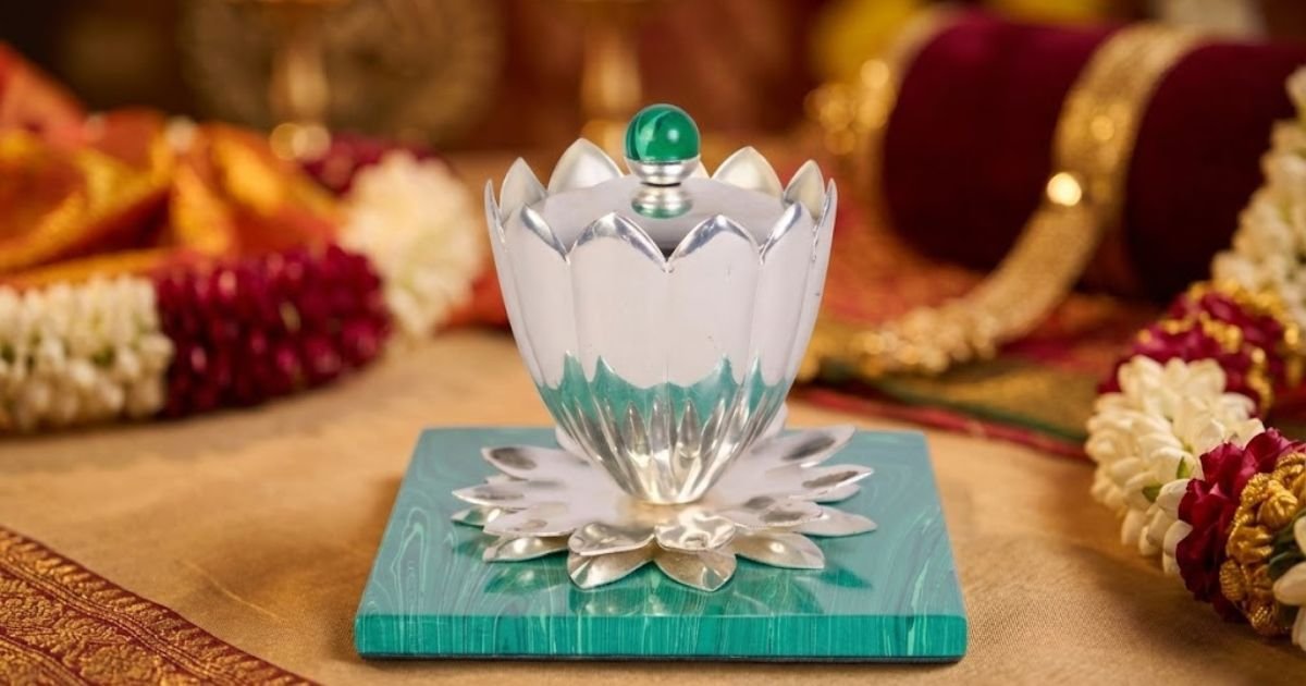 Silver lotus-shaped ceremonial lamp with a green knob sits on a turquoise base, set on gold fabric with flower garlands in the background (Hindu rite).
