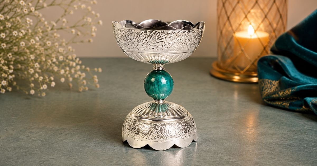 Ornate silver goblet with a teal jewel-like stem on a gray tabletop, candlelit background.