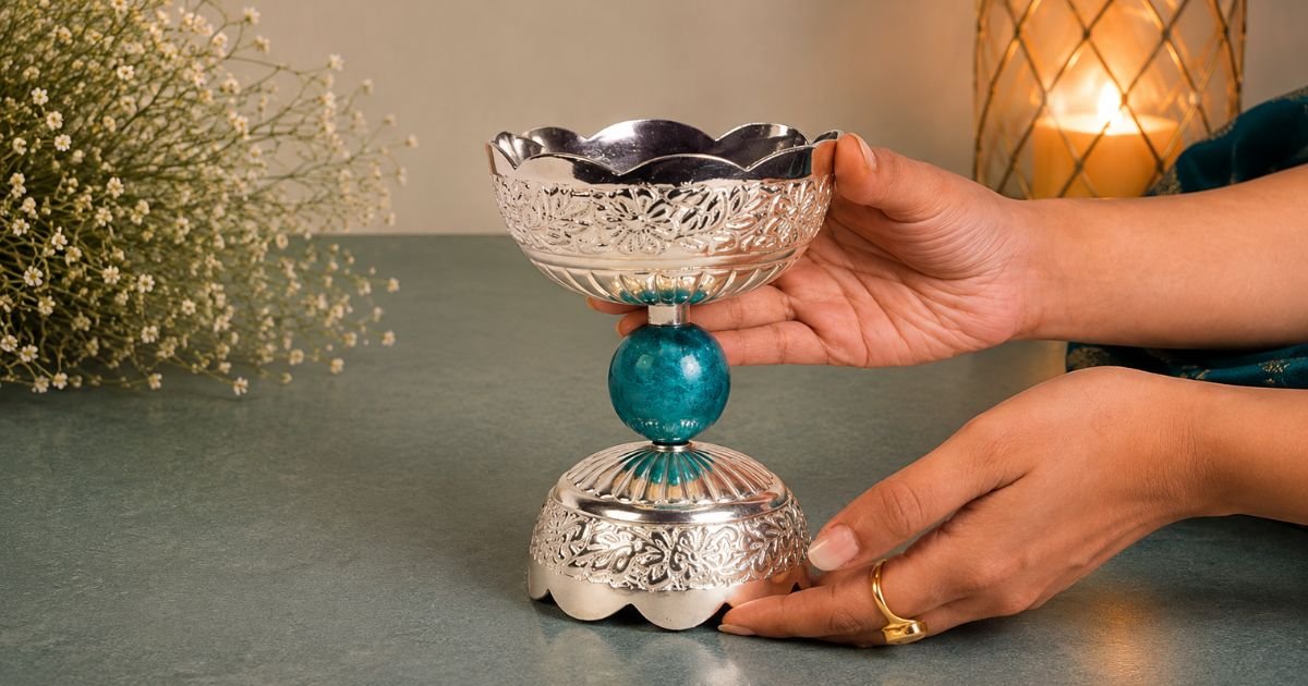 Decorative silver chalice with a turquoise bead stem, being held and stabilized by hands on a teal surface, with a plant and candle in the background.