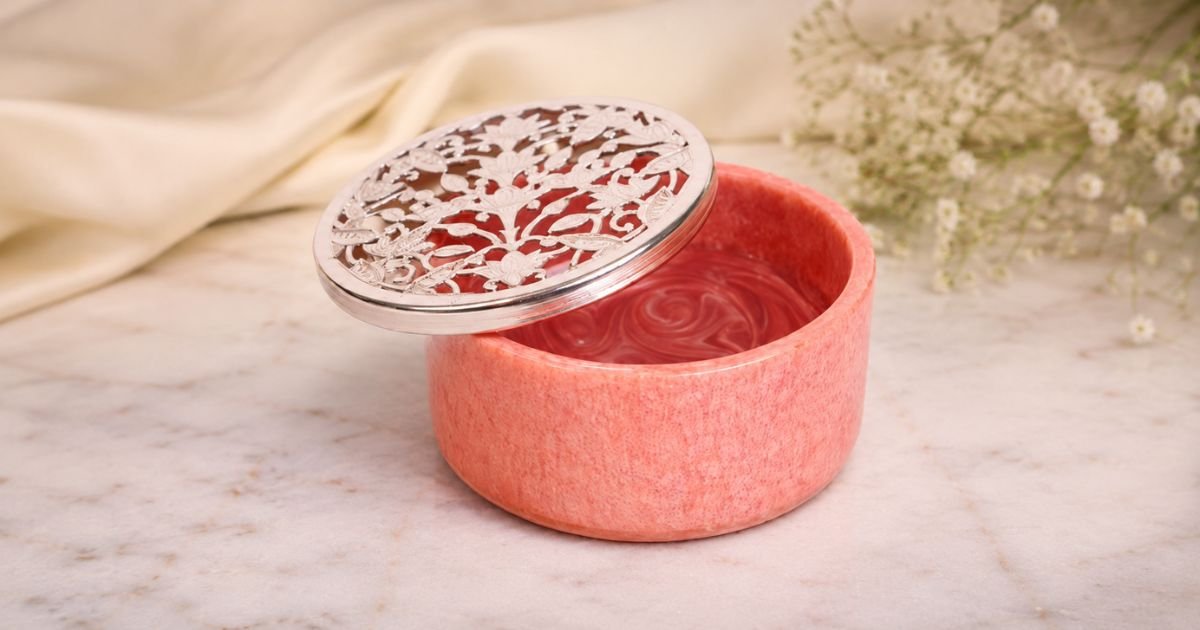 Pink round cosmetic tin with a swirled pink gel inside and an ornate silver lid leaning to the side, set on a marble surface with soft flowers in the background.