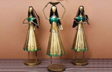 Three gold-toned metal figurines with green accents stand on a striped surface against a peach wall, posing as dancers with arms raised or crossed steadying their balance.