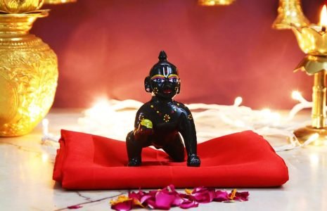 Small black Hanuman statue on a red cloth, part of a lit Hindu altar with golden vessels and candles nearby.
