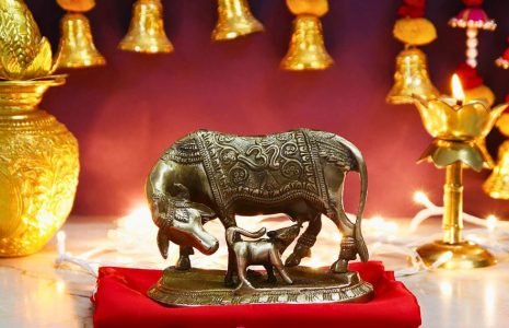 Bronze-toned statue of a cow and calf on a red cloth, displayed on a Hindu altar with lit lamps and gold ornaments in the background.