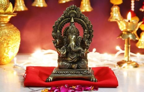 Bronze Ganesha statue seated on a red cushion in a lit shrine with gold decor and petals around it.