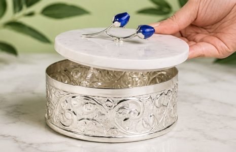 Ornate silver round jewelry box with a white marble lid, hand lifting the lid to reveal intricate scrollwork interior.