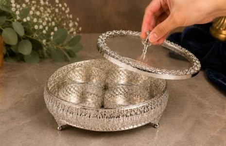 Hand lifting the lid of an ornate silver serving stand that holds four small cups inside.