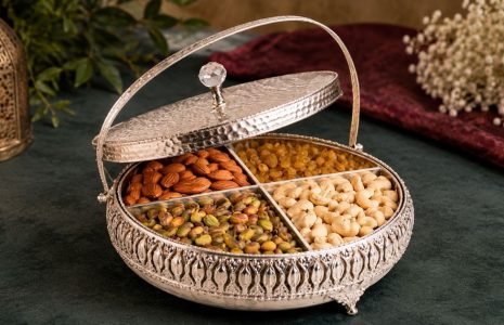 Ornate silver serving box with four compartments filled with almonds, pistachios, cashews, and raisins on a dark table surface.