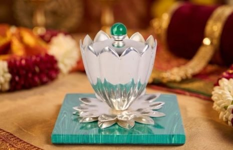 Silver lotus-shaped ceremonial lamp with a green knob sits on a turquoise base, set on gold fabric with flower garlands in the background (Hindu rite).