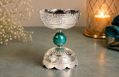 Ornate silver goblet with a teal jewel-like stem on a gray tabletop, candlelit background.
