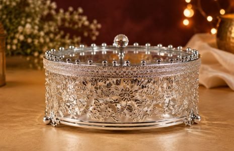 Ornate silver jewelry box with floral lattice and glass lid on a warm gold surface, candlelight in the background.