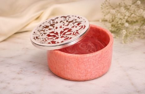 Pink round cosmetic tin with a swirled pink gel inside and an ornate silver lid leaning to the side, set on a marble surface with soft flowers in the background.